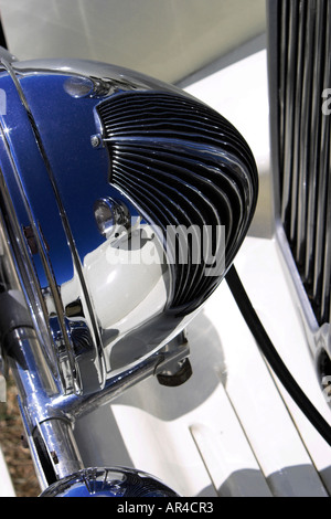 A close up of a headlamp of a white classic automobile and the reflection of the radiator grille curving in the chrome light Stock Photo