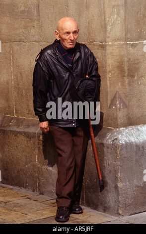 An elderly man leaning against a stone wall while holding a walking cane and a cap in Krakow Poland Stock Photo