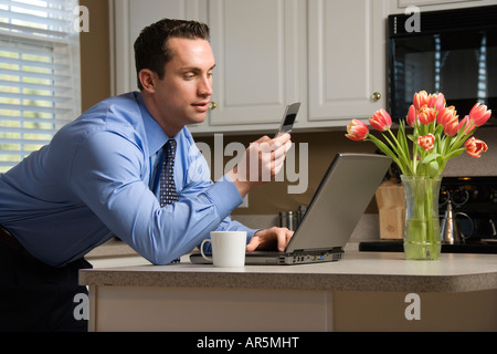 Caucasian man in suit using laptop computer and cellphone in kitchen Stock Photo