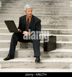 Caucasian middle aged businessman sitting on steps outdoors with laptop and briefcase Stock Photo