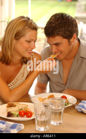 Couple sitting at kitchen table semi profile of female blonde shoulder length hair leaning over to feed crisp to male short Stock Photo