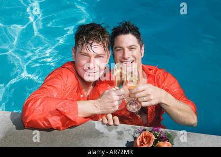 Fully clothed gay couple drinking champagne in swimming pool Stock Photo