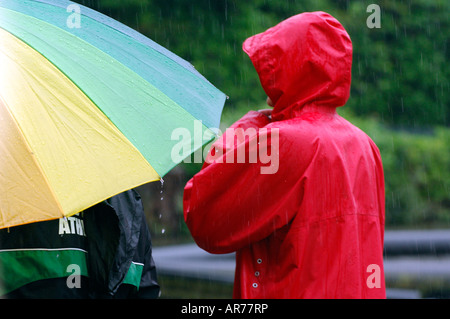 two people walking in the pooring rain with a red jacket with hood up an large green yellow and blue umbrella Stock Photo