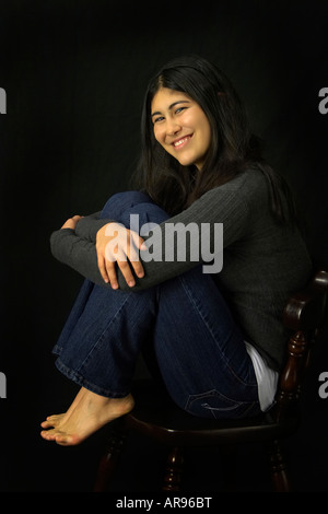 A young Korean American woman with knees pulled up in a chair with a black background Stock Photo