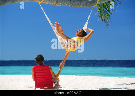 Couple chilling out in hammock and chair under palm tree and blue skies on white sandy beach on Fihalhohi Island in Maldives Stock Photo