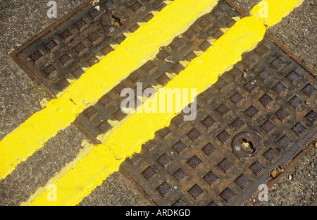 Diagonal detail from above of freshly painted double yellow lines crossing a square metal manhole cover set in tarmac road Stock Photo