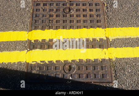 Detail from above of freshly painted double yellow lines crossing a square metal manhole cover set in tarmac road Stock Photo