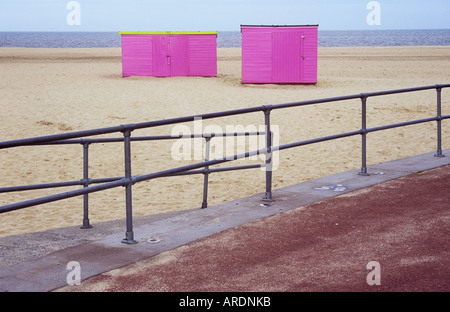 View from sandblown red asphalt promenade across railings to deserted dry white sandy beach with two pink sheds perched on it Stock Photo