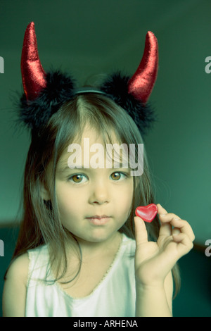 Pretty little girl with devil horns holds heart shaped sweet in hand Stock Photo