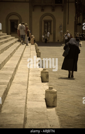 A nun dressed in black walks away from the camera, parallel with the Duomo steps and the bollards. Tourists are in background. Stock Photo