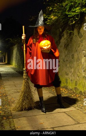 Young woman wearing a red cape and witch hat holding a broomstick and a glowing pumpkin in a solitary alley at night Stock Photo