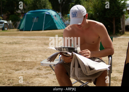 young man sitting with shirt off reading newspaper in the Sun on camp site Stock Photo