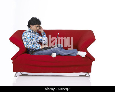 VDA200165 South Asian Indian man sitting on sofa working on laptop and talking on mobile wearing jeans and shirt MR 701 Stock Photo