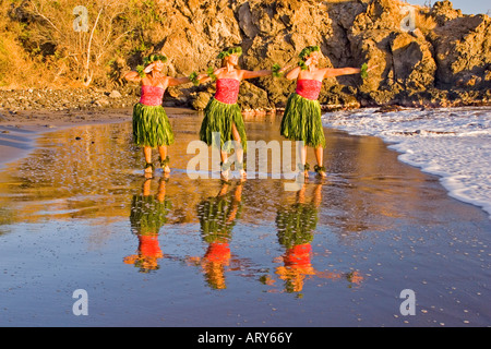 Three hula dancers in ti leaf skirts dance at the water's edge with their reflection at Olowalu, Maui. Stock Photo