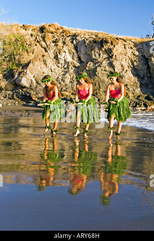 Three hula dancers in ti leaf skirts dance at the water's edge with their reflection at Olowalu, Maui. Stock Photo