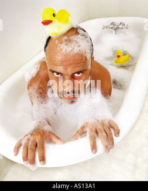 Deranged man hangs on the edge of an old tub in bubble bath and rubber duck on his head Stock Photo