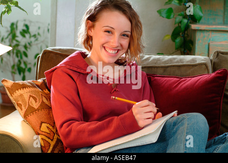 18 year old teenaged girl writing in a notebook on the couch. Perhaps she is working on homework for school or college Stock Photo
