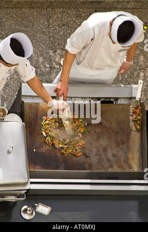 Horizontal aerial close up of two chefs in whites adding ingredients to a stir fry outside on a hotplate Stock Photo