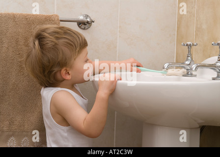 boy toddler twenty two months old cleaning, brushing teeth with toothbrush at basin in bathroom, dressed in white vest, singlet Stock Photo