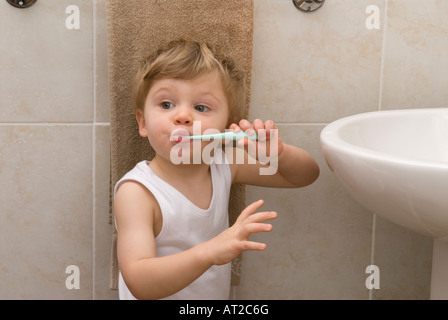 boy toddler twenty two months old cleaning, brushing teeth with toothbrush at basin in bathroom, dressed in white vest, singlet Stock Photo