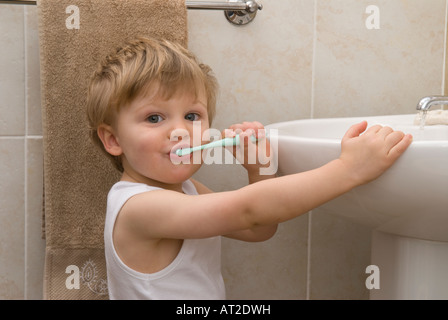 boy toddler twenty two months old cleaning, brushing teeth with toothbrush at basin in bathroom, dressed in white vest, singlet Stock Photo