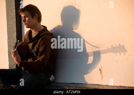 young man, student Aberystwyth University, playing acoustic guitar sunny evening with shadow cast on wall behind him Stock Photo