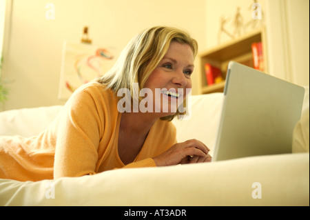 A woman lying on a sofa looking at a laptop, smiling Stock Photo