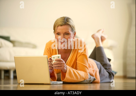 A woman lying on the floor holding a cup and looking at a laptop Stock Photo