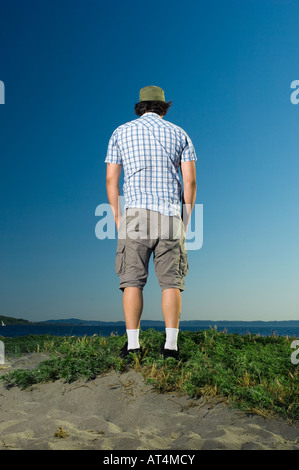 Man in thirties wearing shorts and a hat standing on a grassy hill and overlooking the water. Stock Photo