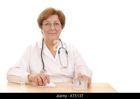 Senior family doctor with stethoscope sitting behind a desk with fingers put on alarm clock Stock Photo