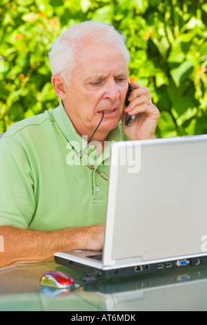 A mature man looking frustrated unhappy while talking on his cell phone and working on a laptop computer Stock Photo