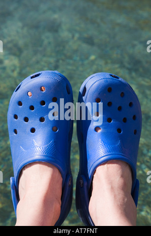 Pair of person's feet wearing blue Crocs plastic sandals against clear sea water Stock Photo