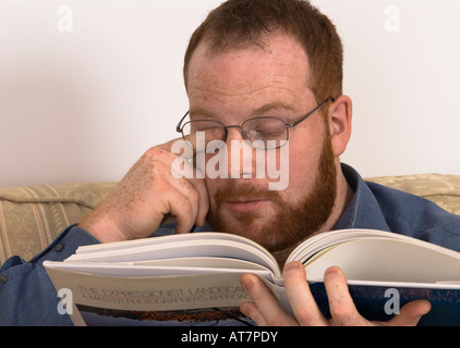 young man with poor eyesight wearing glasses rubbing his strained eye with finger trying to read book, incorrect spectacles Stock Photo