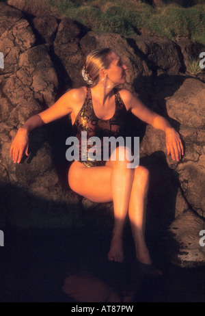 A beautiful woman in a black swimsuit poses in natural outdoor rock hot tub. Stock Photo