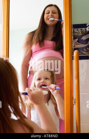 Pregnant woman brushing teeth in bathroom mirror dressed in pink pyjamas with daughter Stock Photo