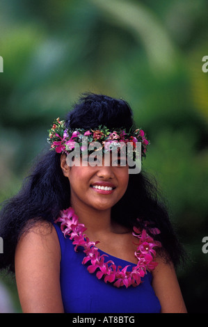 A beautiful young Samoan woman wearing a pink lei and haku lei smiles against a background of muted green foliage. Stock Photo