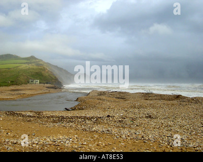 Stormy day with large waves and spray breaking at Charmouth on the Dorest coast A small river estuary is flowing into the sea across a stone pebble and sand beach Stock Photo