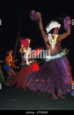 Two colorful female Tahitian dancers shake to the beat while a male dancer wearing shorts and a lei performs in the background Stock Photo