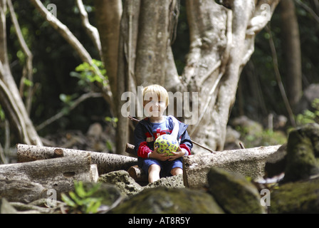 small blonde boy holds a ball while sitting on a log beside a large tree at Limahuli Gardens, on Kauai's majestic north shore. Stock Photo