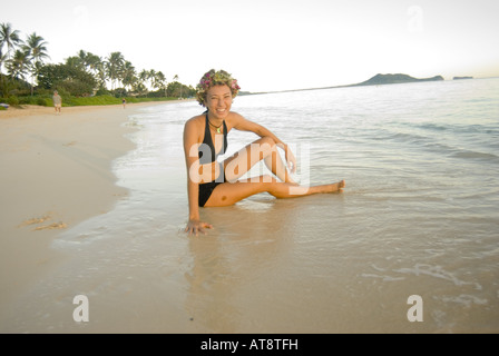 Beautiful young woman wearing black swimsuit sits in water by shore at world famous Lanikai beach on windward side of Oahu Stock Photo