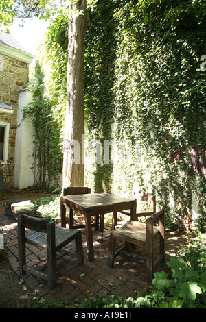 Small heavy square wooden table with four heavy wooden chairs around it on small brick patio area with tall ivy covered wall Stock Photo