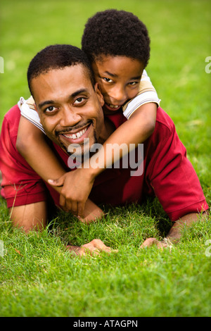 Father lying in grass smiling as son climbs on his back and hugs his neck Stock Photo
