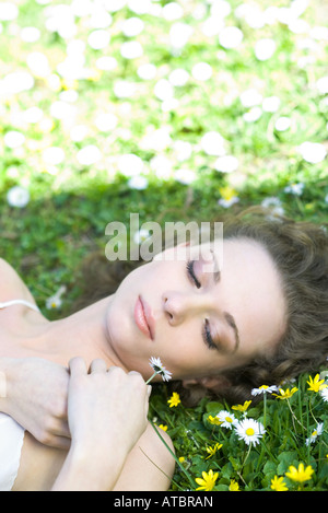 Young woman lying in on back on the ground, holding flower, eyes half-opened Stock Photo