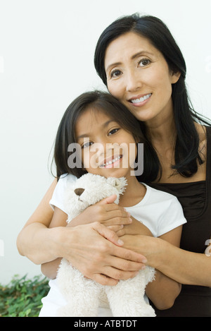 Mother and daughter embracing teddy bear together, both smiling at camera, portrait Stock Photo