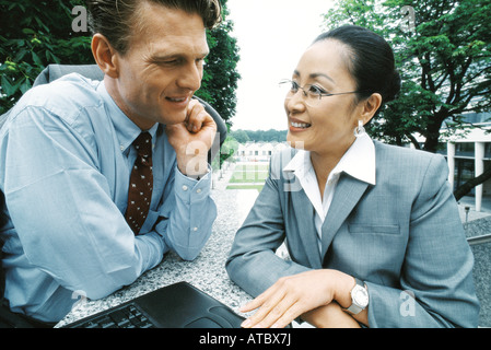 Business partners sitting outdoors, smiling, woman using laptop computer Stock Photo