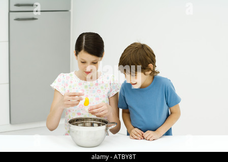 Two young siblings cooking together, girl cracking egg, both looking down Stock Photo