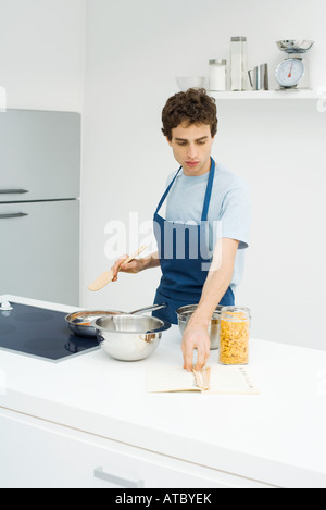 Young man cooking in kitchen, reading recipe from book Stock Photo