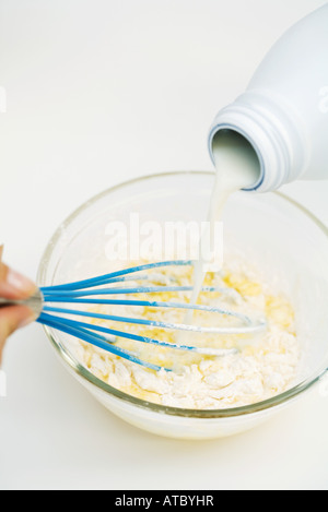 Person pouring milk into mixing bowl, holding whisk, cropped view Stock Photo