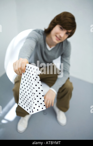 Young man sitting in chair, holding gift bag toward camera, focus on foreground Stock Photo