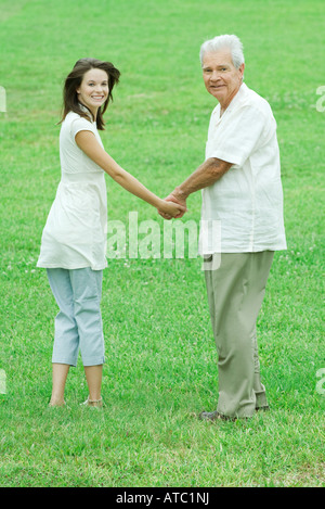 Grandfather and teenage granddaughter holding hands, both smiling over shoulders at camera Stock Photo